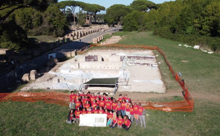 Gruppo di partecipanti alla campagna di scavo 2025 nell'area R. V, Is. XIII di Ostia antica, davanti al cantiere archeologico ripreso dall'alto.